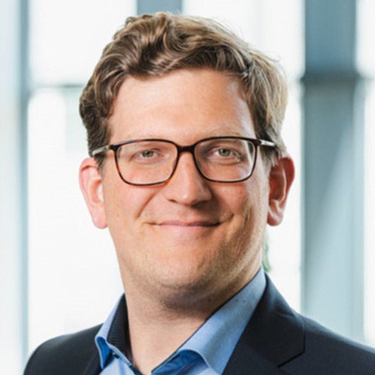 Portrait Pressesprecher Marc Friedrich. Mann trägt Brille und Anzug; lächelt; steht vor hellen Fenstern.