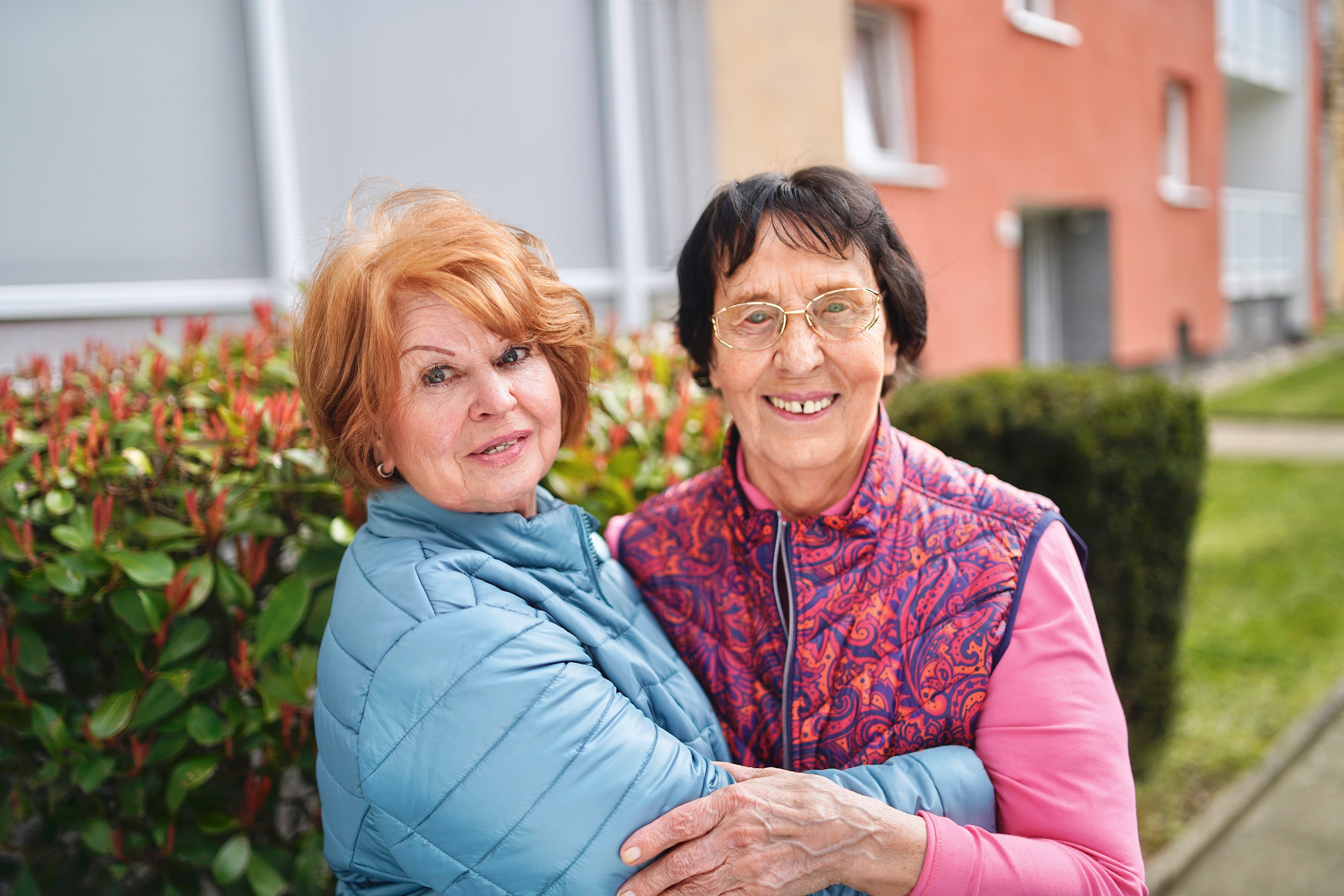 Zwei ältere Frauen umarmen sich vor einem Wohnhaus.