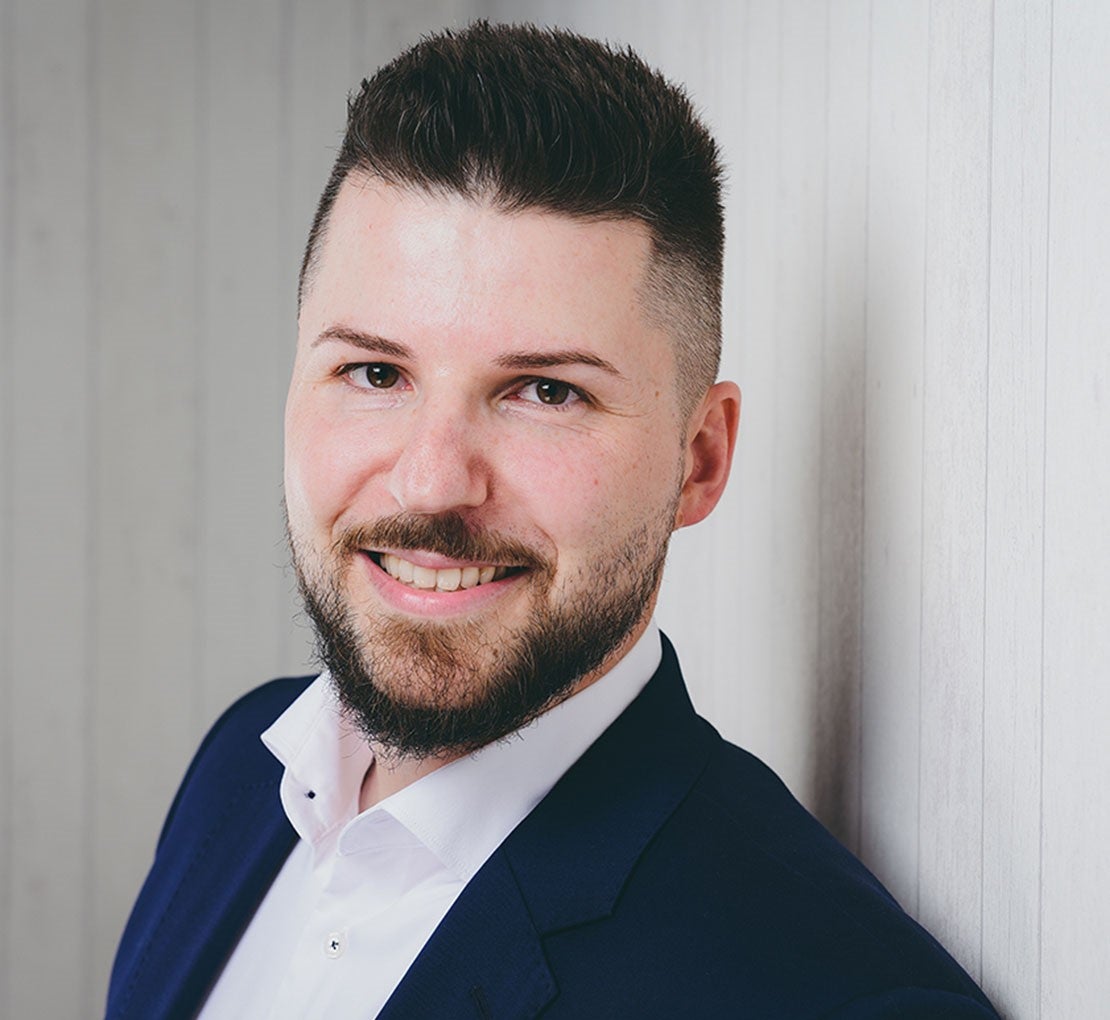 Portrait Andreas Knaust. Mann lächelt; trägt Anzug; steht vor heller Holzwand.
