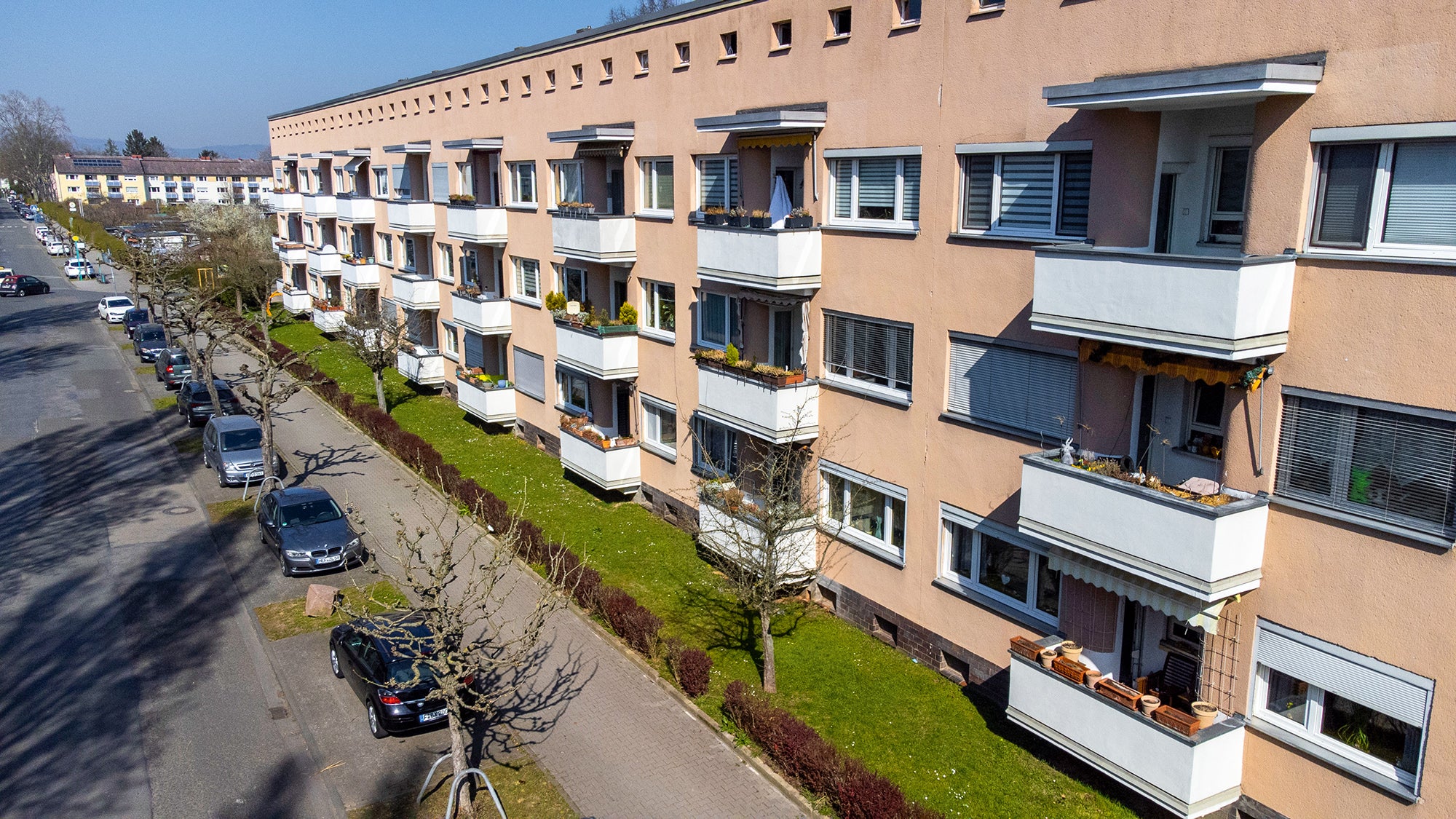 Wohnblock mit Balkonen, Autos parken auf der Straße, Bäume ohne Blätter.