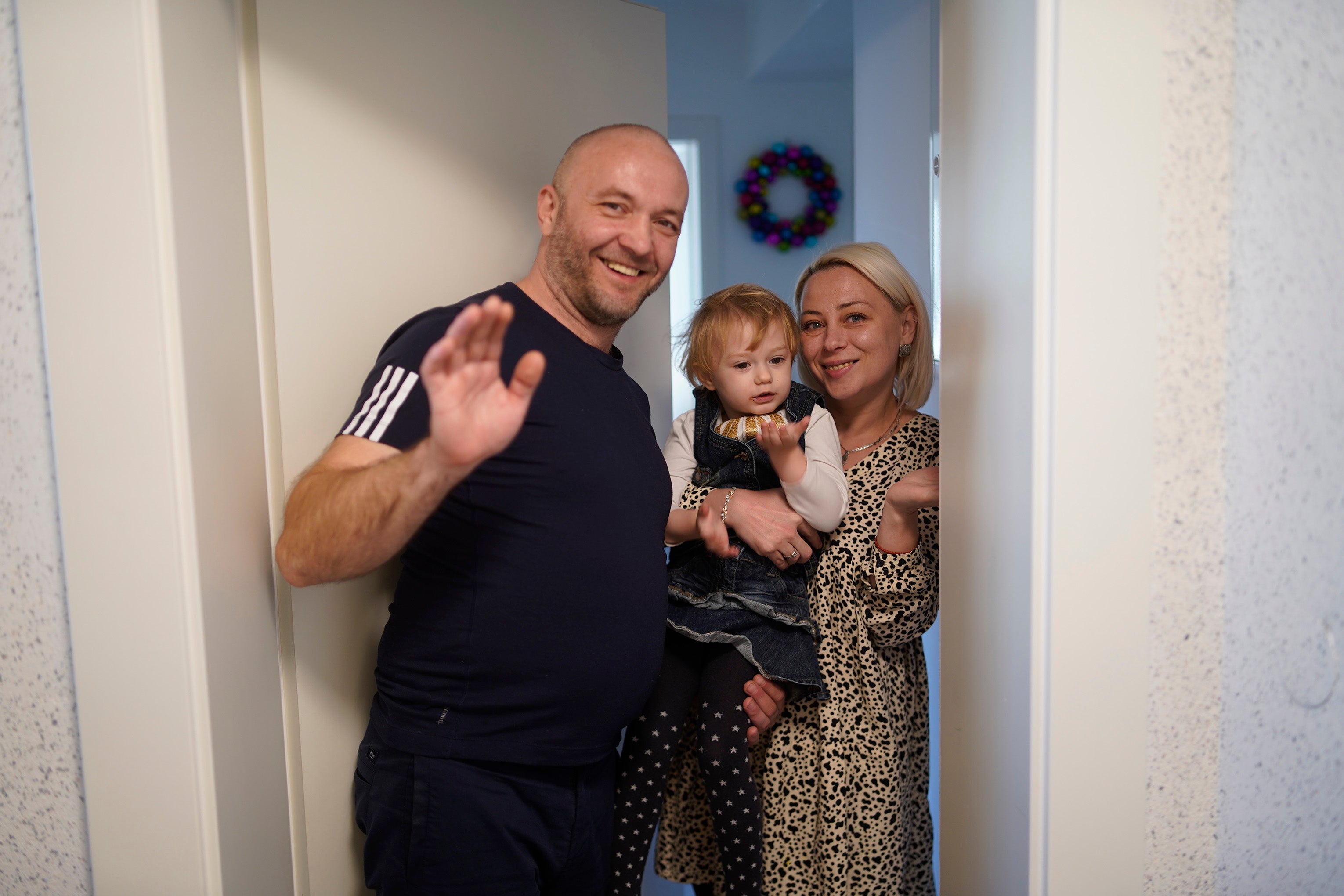 Familie winkt lächelnd an einer Tür; Kranz im Flur zu erkennen.