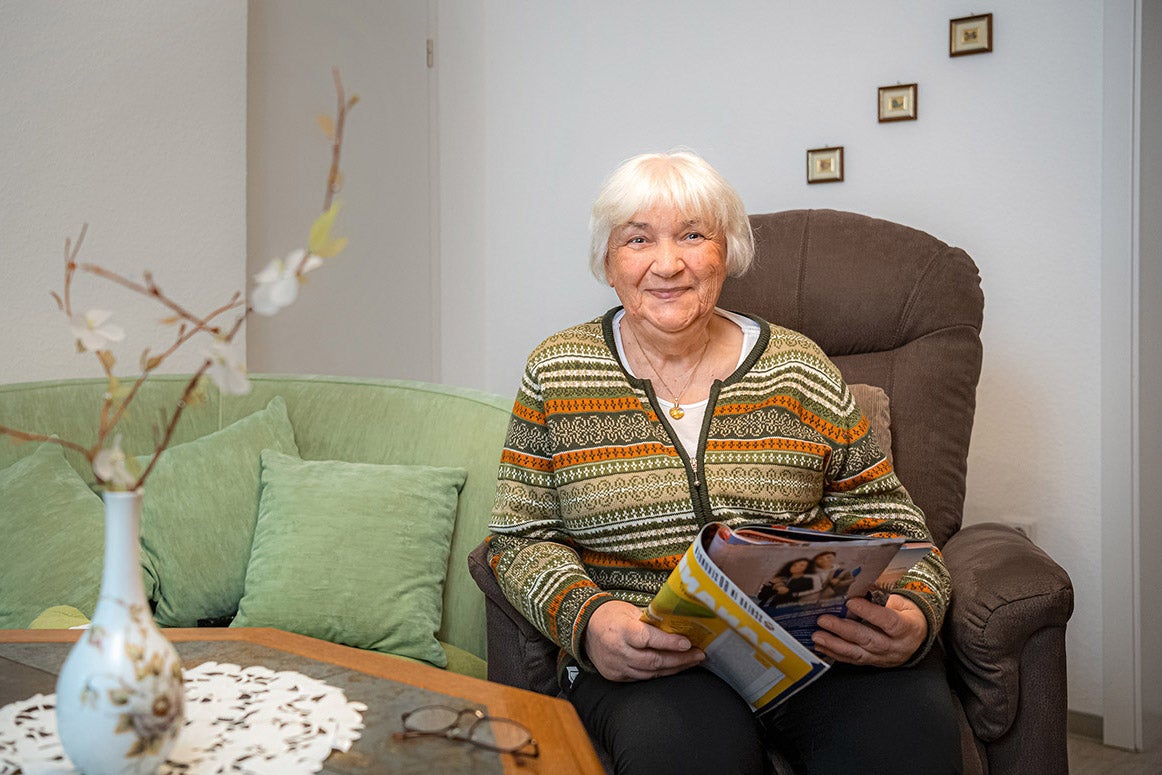 Frau sitzt lächelnd in einem Sessel, hält ein Magazin im Wohnzimmer.