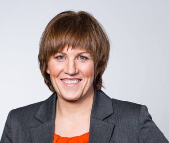 Portrait Presseleitung Nina Henckel. Frau lächelt und trägt grauen Anzug; neutraler Hintergrund.