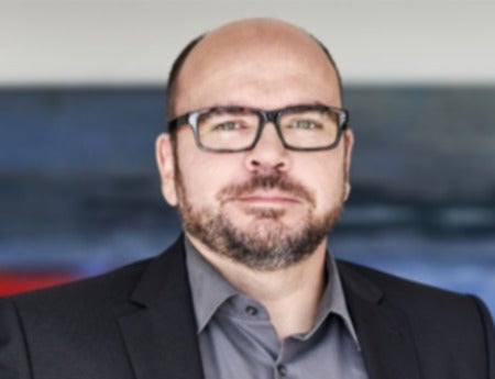 Portrait Pressesprecher Olaf Frei. Mann mit Bart trägt Brille und dunklen Anzug, steht vor verschwommenem Hintergrund.