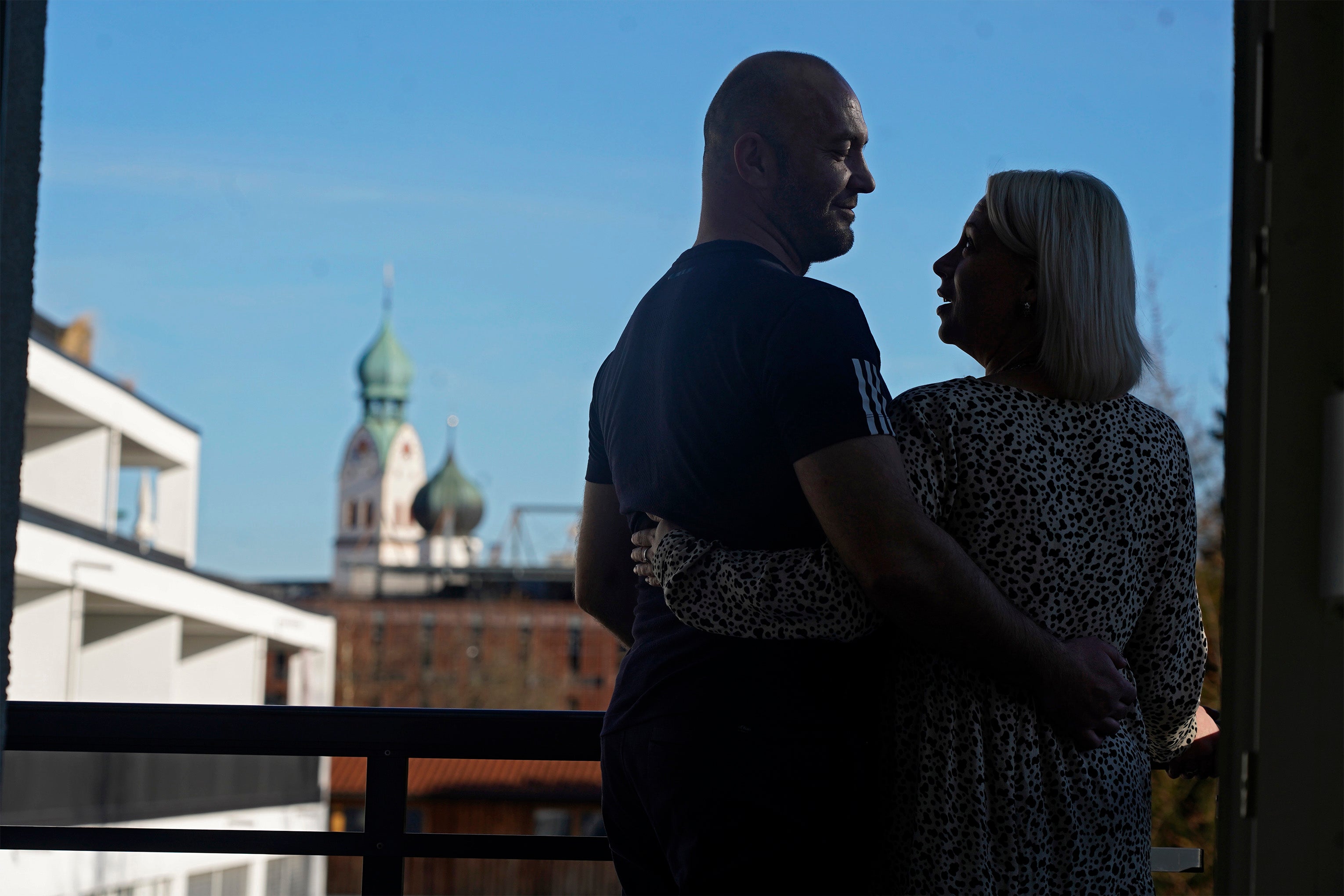 Paar umarmt sich auf einem Balkon mit einem Kirchturm im Hintergrund.