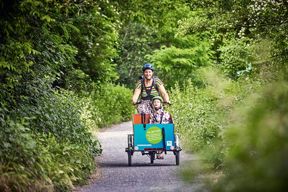 Frau fährt Lastenrad mit Kind durch bewaldeten Pfad.