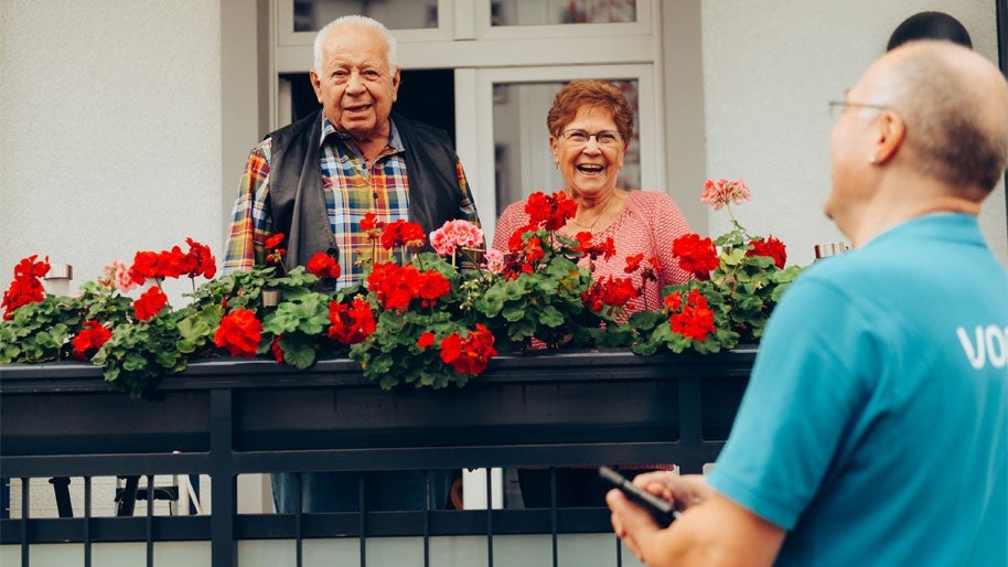 Älteres Paar lächelt vom Balkon aus, Vonovia Mitarbeiter unscharf im Vordergrund.