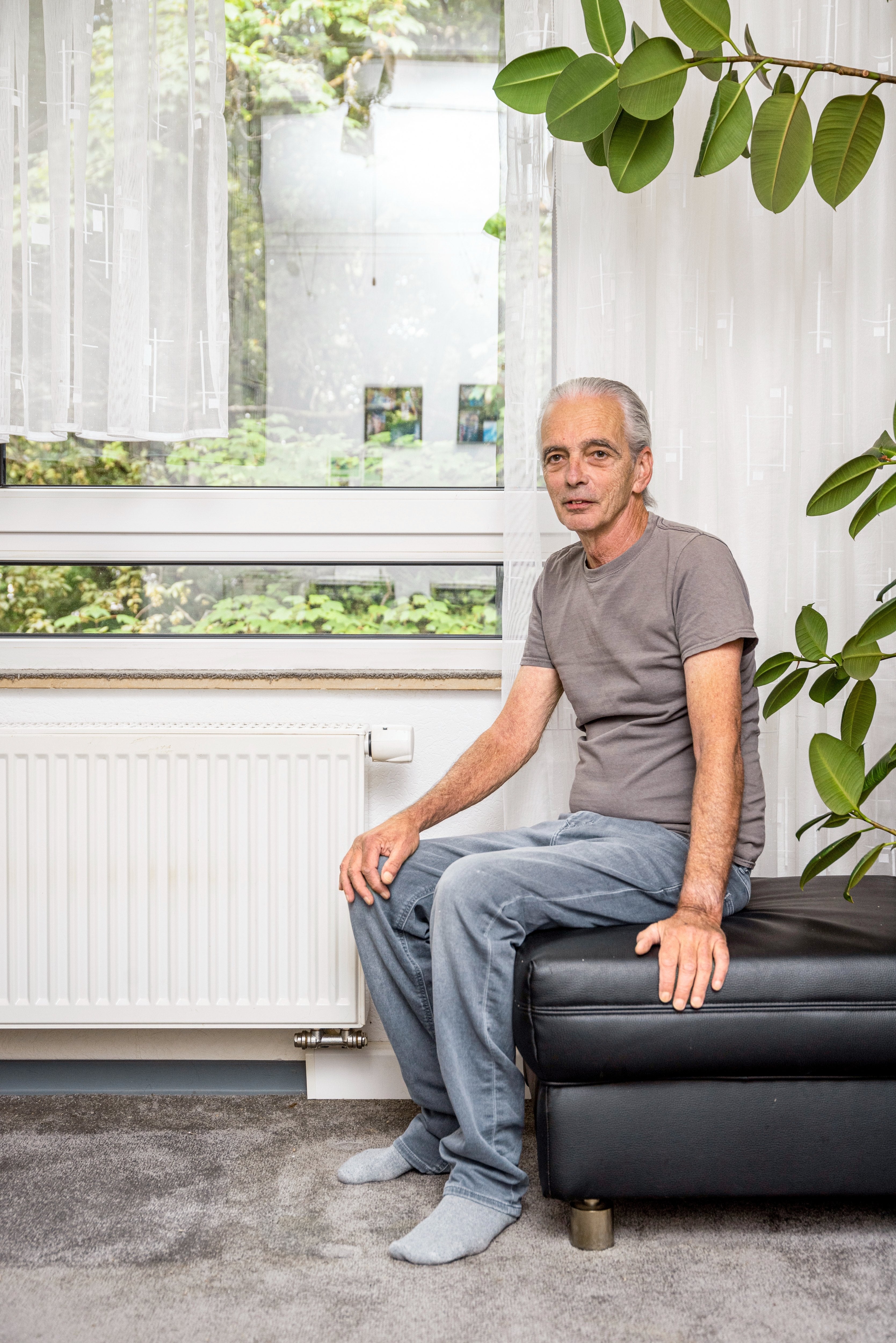 Heinz Dietrich aus Wiesbaden. Mann sitzt auf Bank in hellem Wohnzimmer mit großen Fenstern vor einer Heizung.