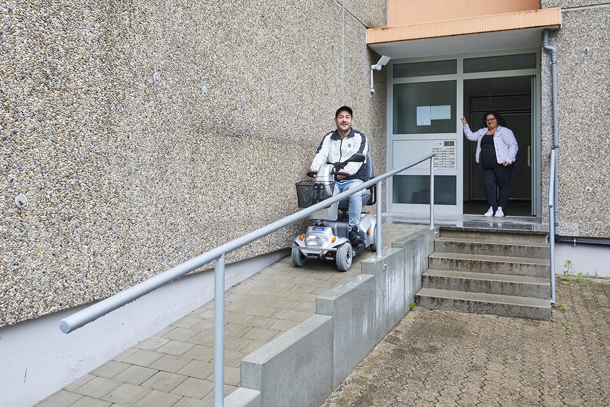 Ein Mann fährt auf einem Elektromobil eine Rampe am Eingang seines Wohnhaues herunter. In der Tür steht eine Frau und schaut zu. Beide freuen sich über die Rampe.