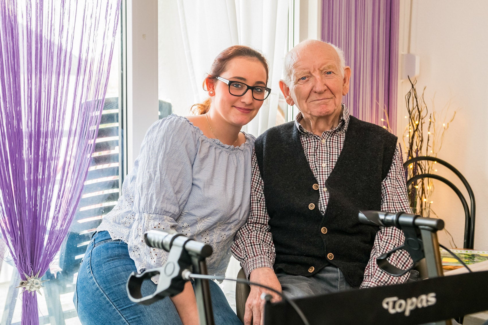 Eine Frau und ein älterer Mann sitzen im Wohnzimmer hinter einem Rollator.